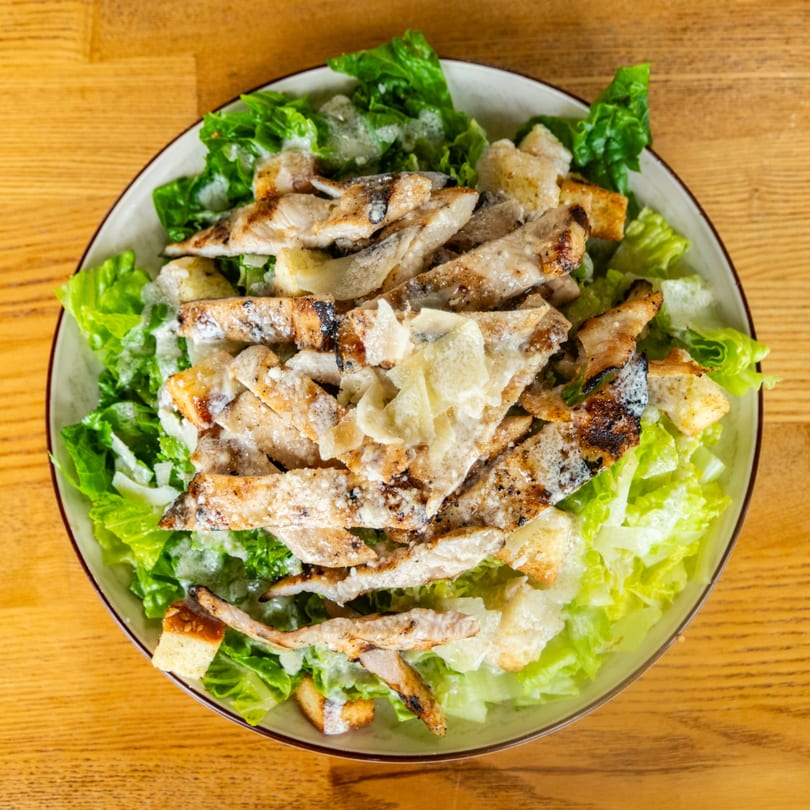 A Caesar salad with grilled chicken, croutons, and parmesan cheese in a bowl on a wooden table.