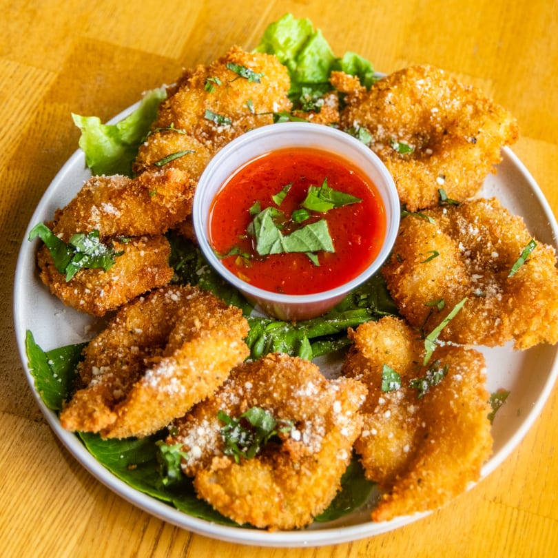 Plate of golden fried ravioli with red dipping sauce, garnished with parmesan and basil.
