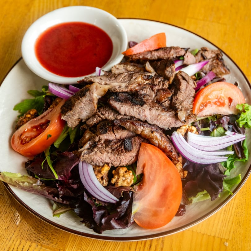 Plate of grilled steak salad with tomatoes, red onions, walnuts, and a side of red dressing.
