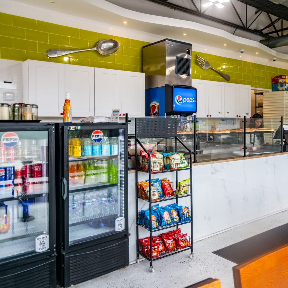 Restaurant counter with green tiles, Pepsi coolers, snack rack, pizza prep, and large decorative spoon and fork.