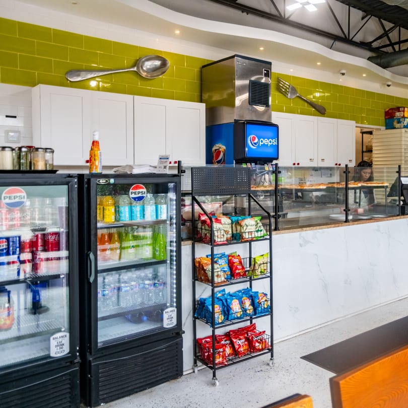 Restaurant counter with green tiles, Pepsi coolers, snack rack, pizza prep, and large decorative spoon and fork.