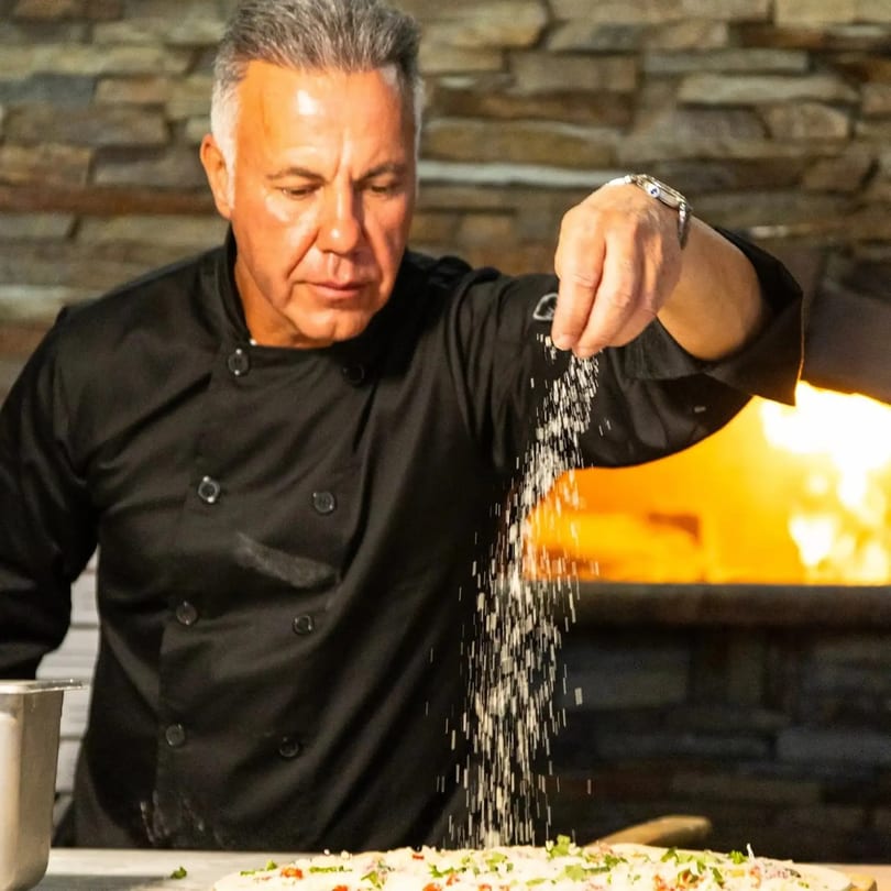 Chef sprinkles cheese onto a pizza in front of a blazing wood-fired oven.
