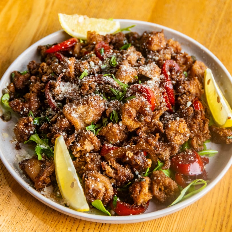 Crispy fried calamari with red peppers, parmesan, fresh herbs, and lemon wedges on a white plate.