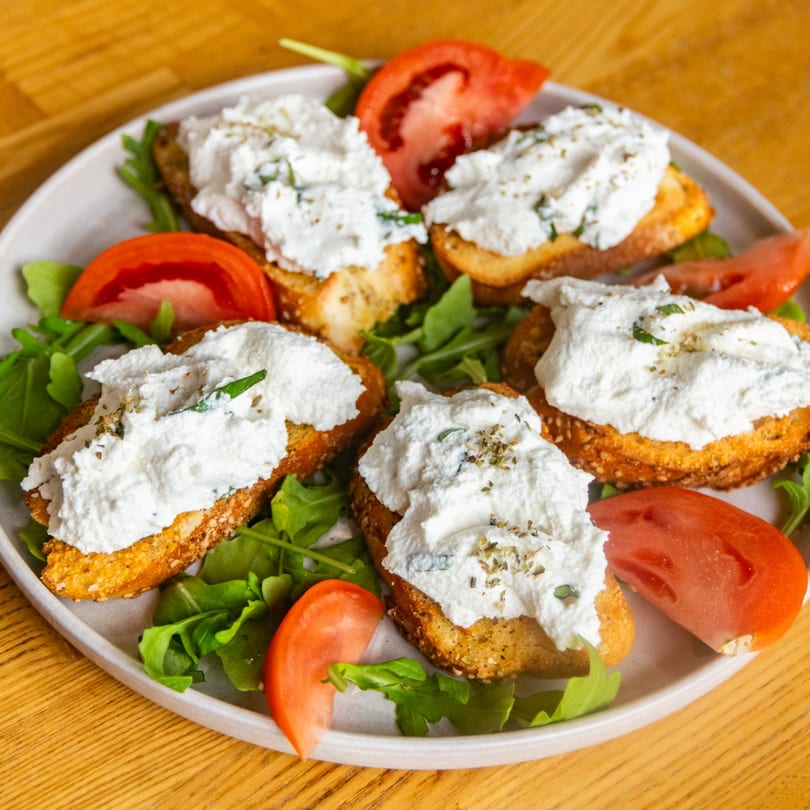 Plate of toasted bread topped with creamy white cheese, herbs, and tomato slices on arugula.