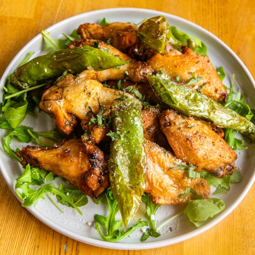 Crispy golden chicken wings with shishito peppers, arugula, and grated cheese on a white plate.