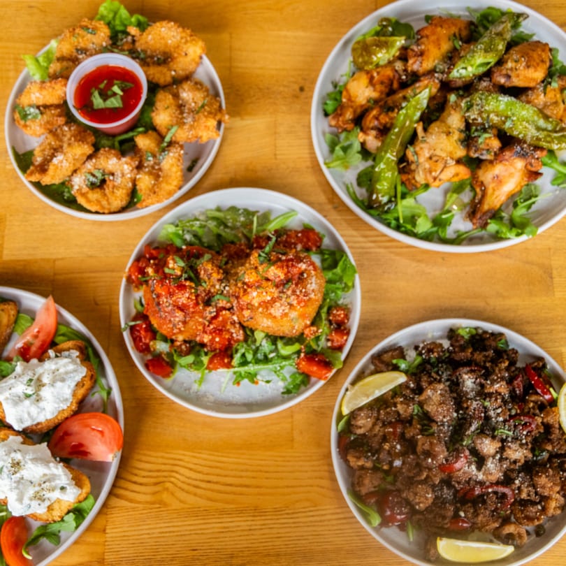 Overhead view of five plates of diverse appetizers, including fried seafood, chicken, and bruschetta, on a wooden table.