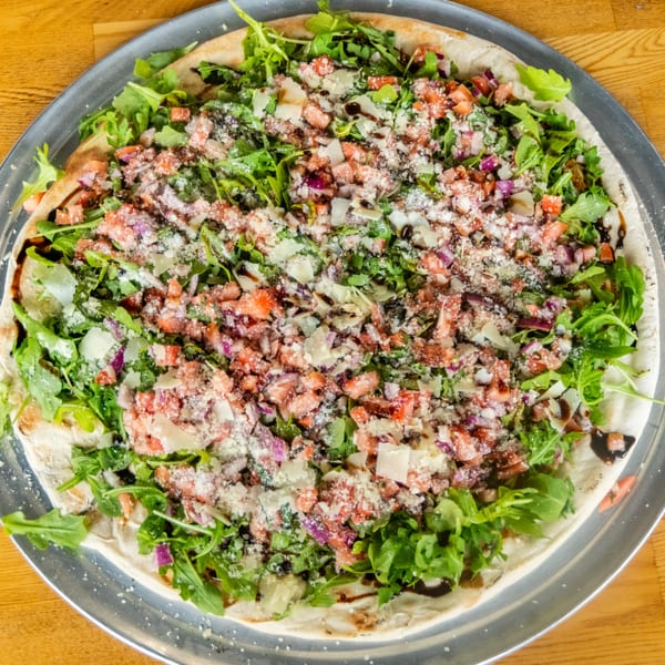 Flatbread pizza topped with fresh arugula, diced tomatoes, red onions, parmesan, and balsamic glaze on a metal pan.