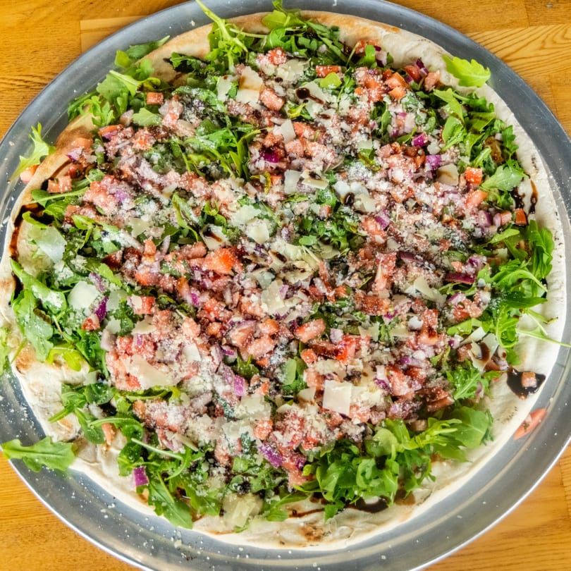 Flatbread pizza topped with fresh arugula, diced tomatoes, red onions, parmesan, and balsamic glaze on a metal pan.