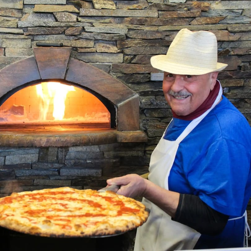 Smiling pizzaiolo holding a fresh pizza in front of a blazing stone oven.