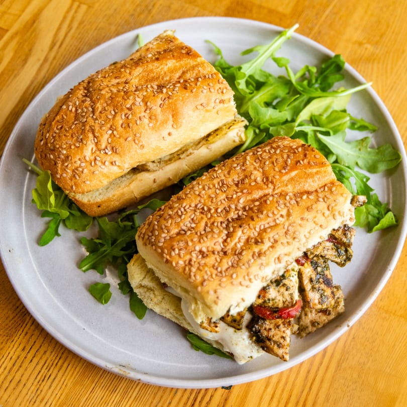 Two grilled chicken sandwiches with melted cheese and arugula on sesame seed rolls.