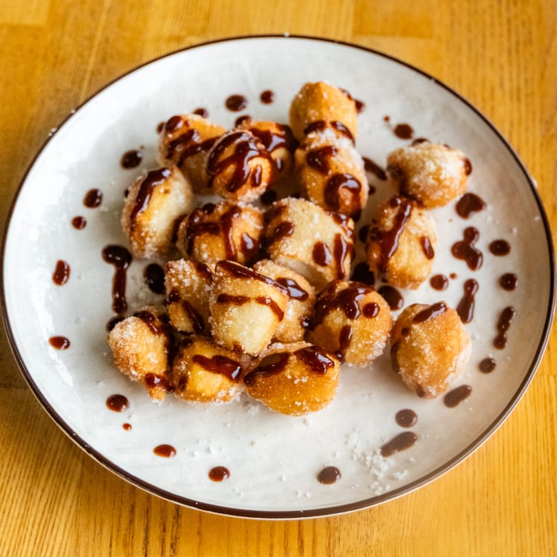 Sugar-dusted fried dough pieces drizzled with chocolate sauce on a white plate.