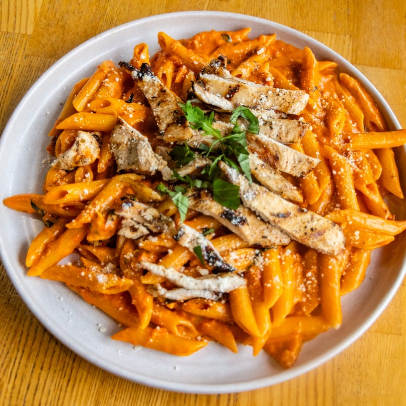 Penne pasta with grilled chicken, creamy tomato sauce, Parmesan, and fresh basil on a white plate.
