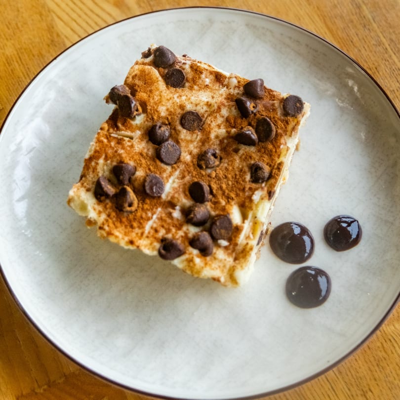 Square dessert slice with chocolate chips and cinnamon powder on a plate with chocolate drops.