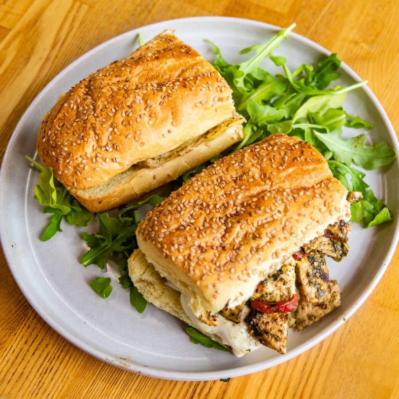 Two sesame seed baguette halves filled with grilled chicken, cheese, and arugula on a grey plate.