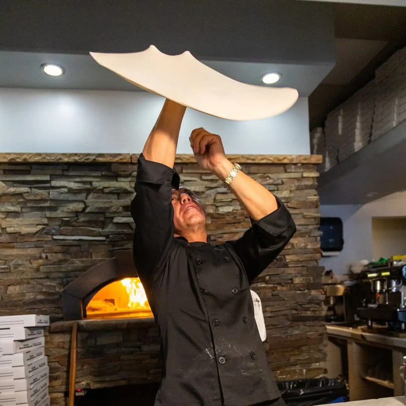 Chef in black uniform expertly tosses pizza dough high above a wood-fired oven.