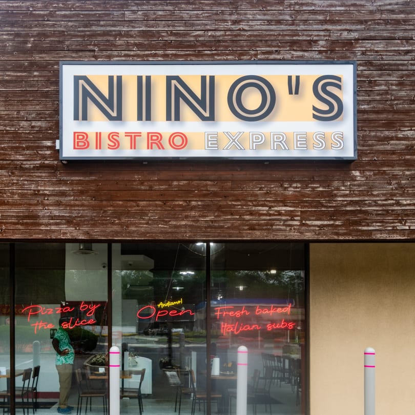 Nino's Bistro Express storefront with a large sign, wooden facade, and neon window signs advertising pizza and subs.
