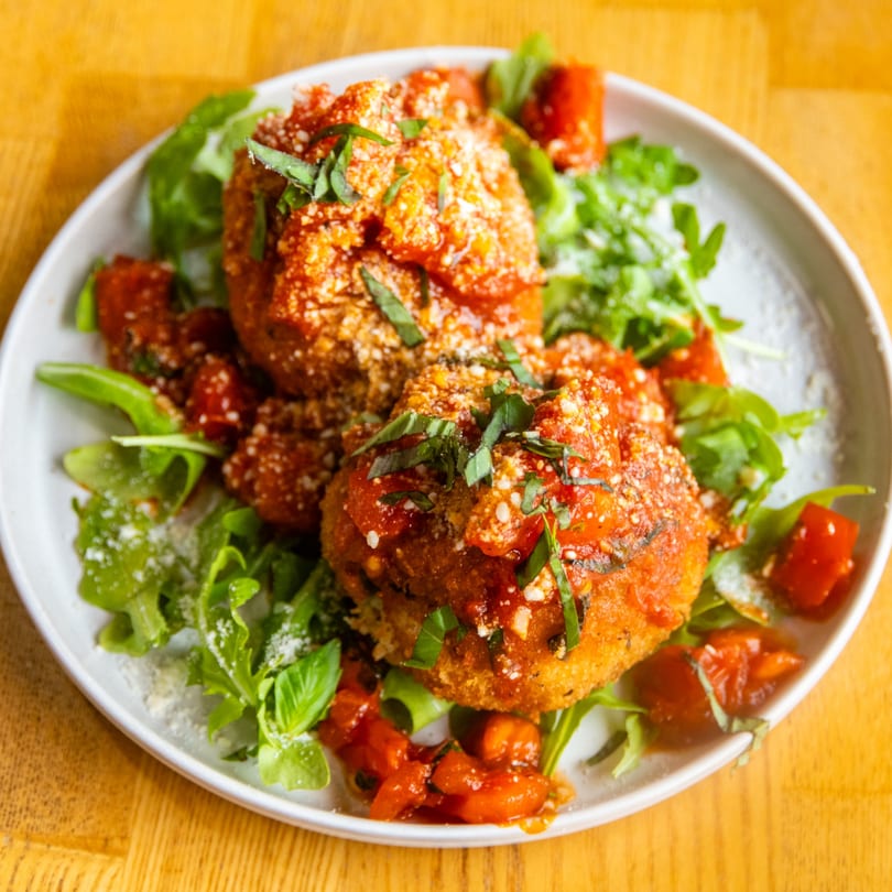 Two fried arancini with tomato sauce, parmesan, and basil on a bed of arugula and diced tomatoes.