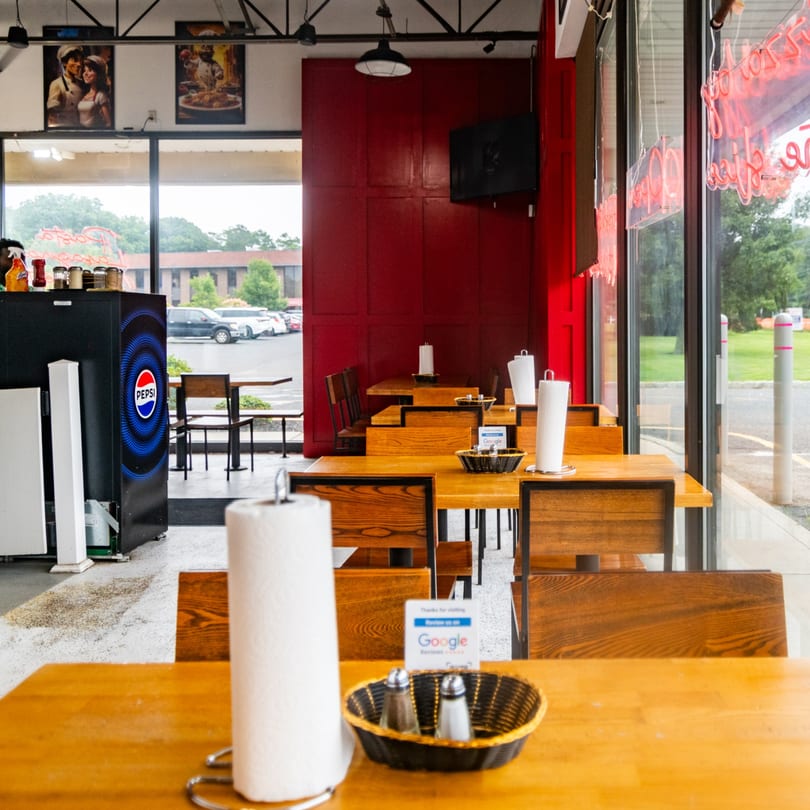 Bright restaurant interior with wooden tables, red wall, Pepsi fridge, and large windows overlooking a parking lot.