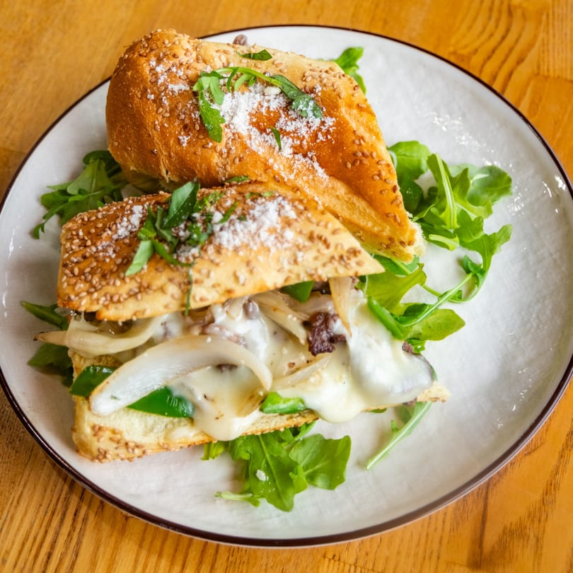 A hearty sandwich with melted cheese, onions, green peppers, and arugula on a sesame seed bun.