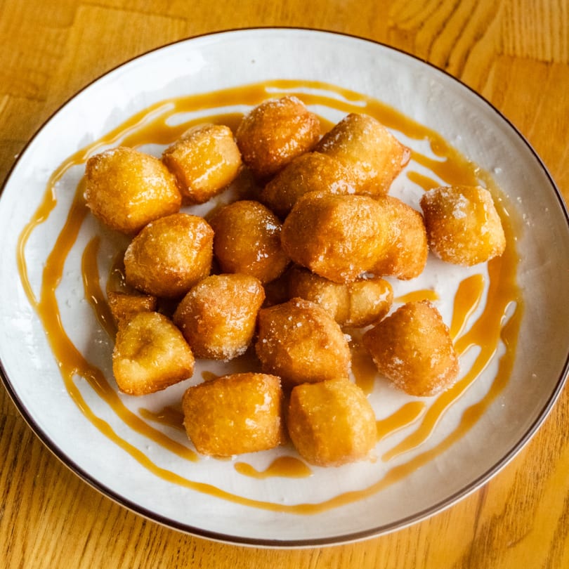 A plate of golden fried dough bites, sugared and drizzled with caramel, on a wooden table.